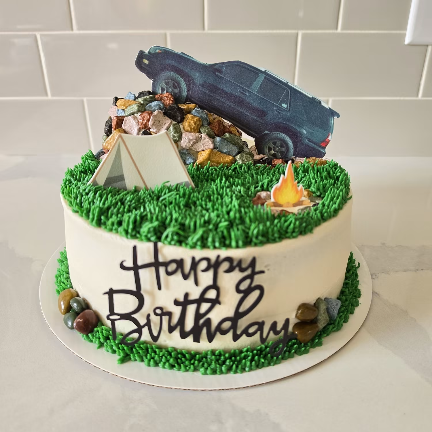 A birthday cake with piped green grass, a fondant tent, campfire and toy SUV on top